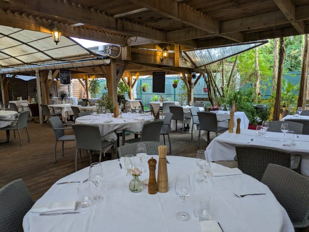 Terrasse Restaurant Élégante: Dîner en Plein Air & Verdure Terrasse de restaurant couverte en bois, tables dressées en blanc, chaises en osier. Verdure luxuriante, lanternes et menu COTE 29.5€.