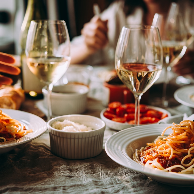 Table de dîner conviviale avec assiettes de spaghetti, verres de vin blanc, parmesan et tomates. Mains de convives au fond.