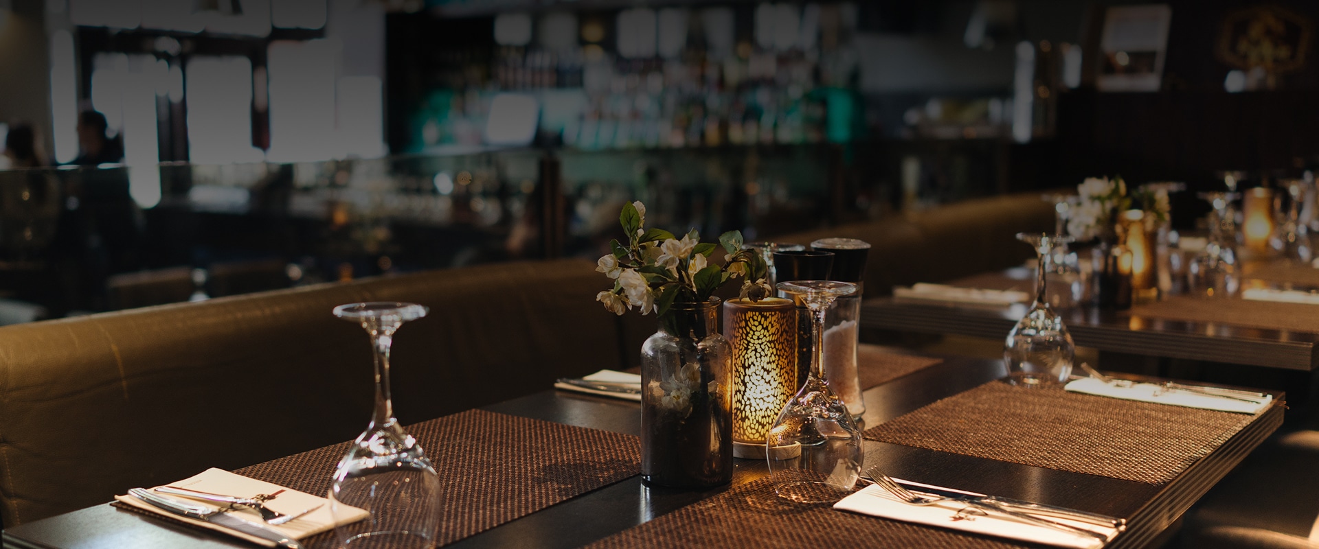 Table de restaurant dressée avec verres, couverts, fleurs et bougie. Arrière-plan flou d'un bar animé. Ambiance chaleureuse.