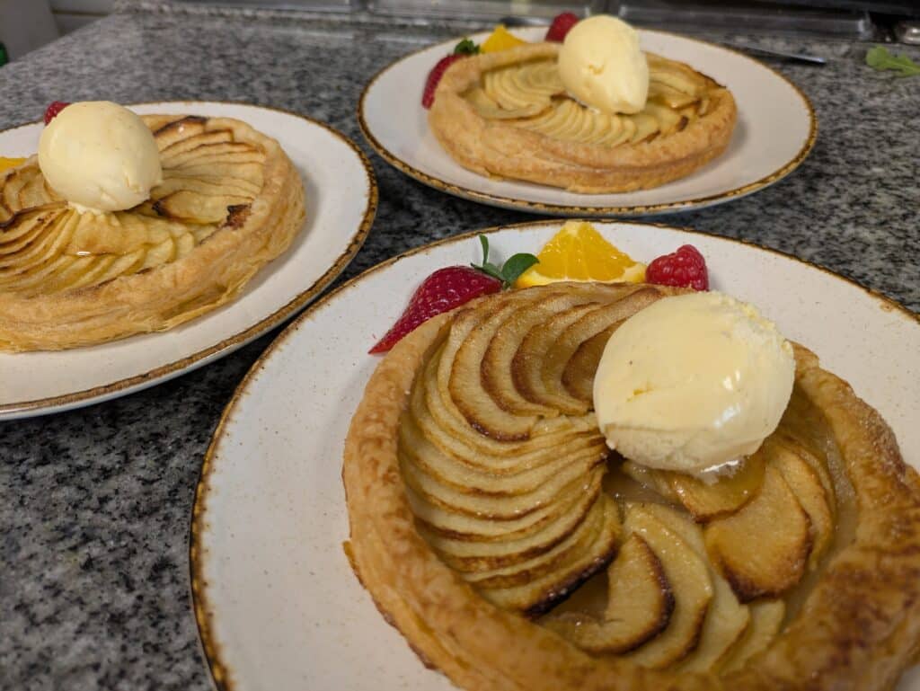 Trois tartes aux pommes individuelles sur assiettes blanches, garnies de tranches de pommes, une boule de glace vanille, fraises et framboises. Comptoir en granit.