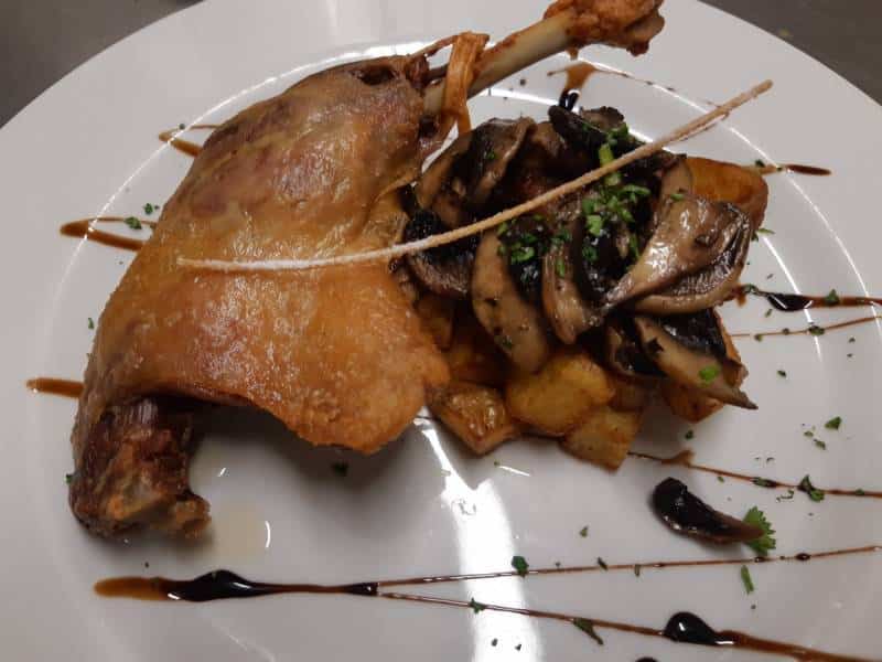 Canard confit, pommes de terre rissolées & champignons Cuisse de canard confite dorée avec peau croustillante, accompagnée de pommes de terre rissolées et champignons de Paris poêlés, sauce au jus.