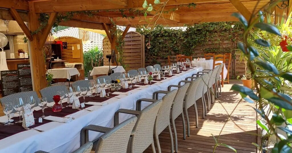 Terrasse de restaurant: table prête sous pergola bois Longue table dressée sous une pergola en bois pour un repas extérieur. Nappe blanche, chemin bordeaux, verres. Panneaux 'MENU CARTE'.