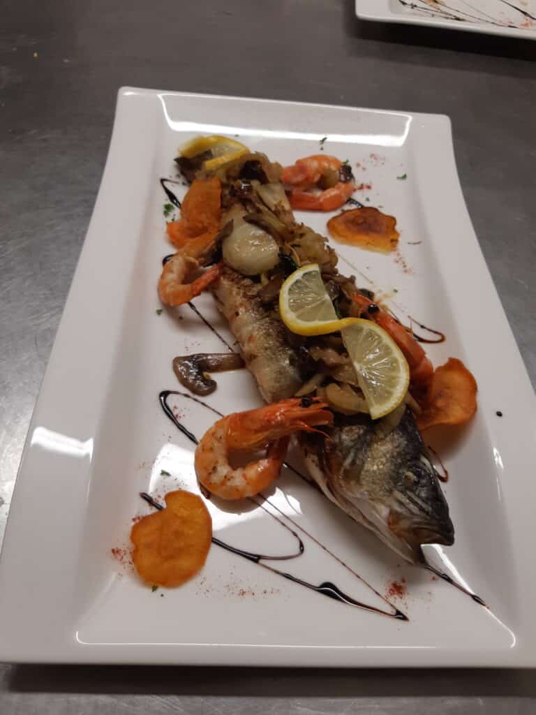Bar entier grillé accompagné de crevettes, citrons et chips, nappé de glaçage balsamique sur assiette blanche.